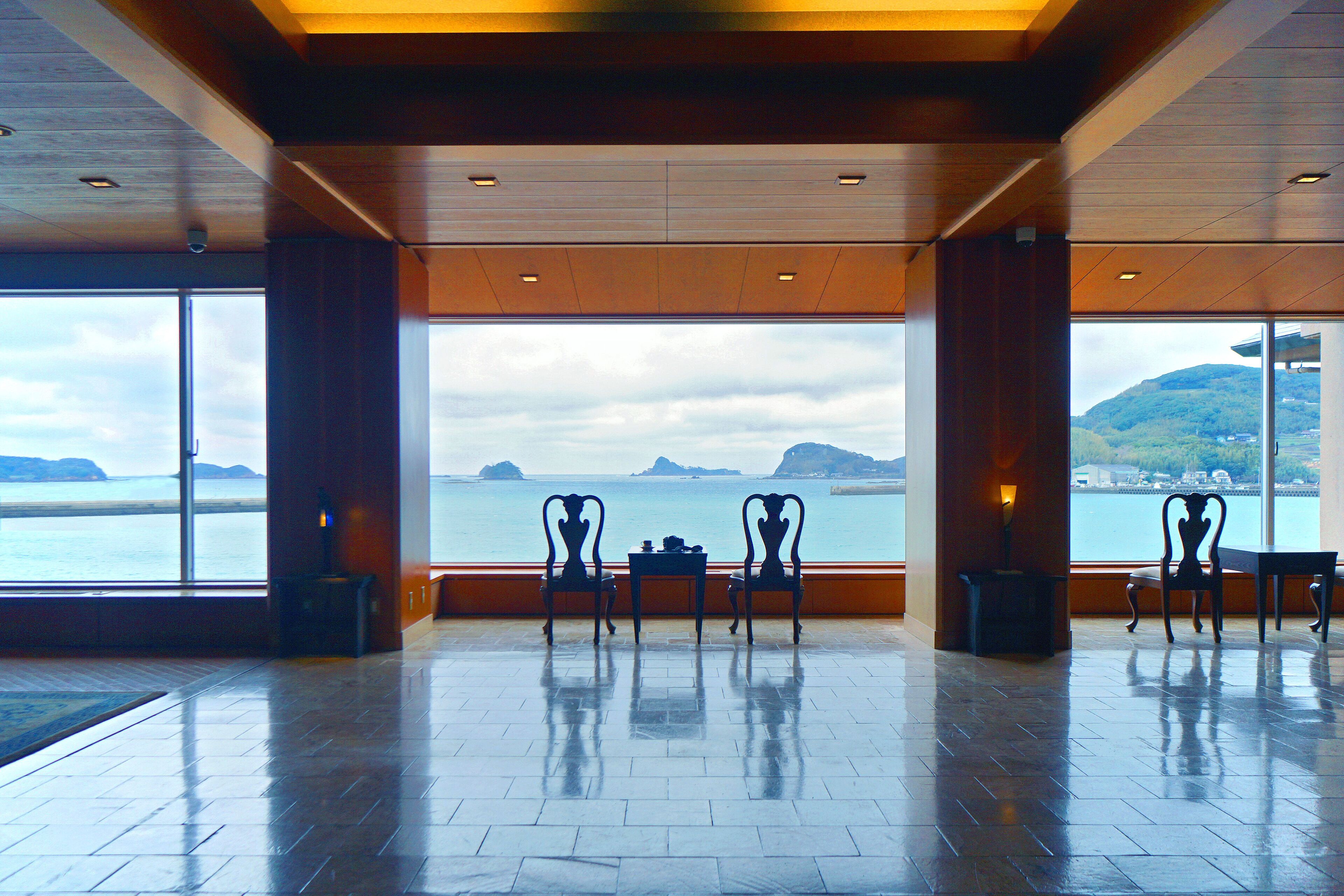 Un amplio espacio interior con un gran ventanal que ofrece vistas al mar y montañas distantes. Hay varias sillas y una pequeña mesa en el centro.