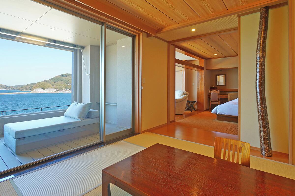 Een kamer met een houten tafel, een bank met kussen, een bed en uitzicht op zee.
