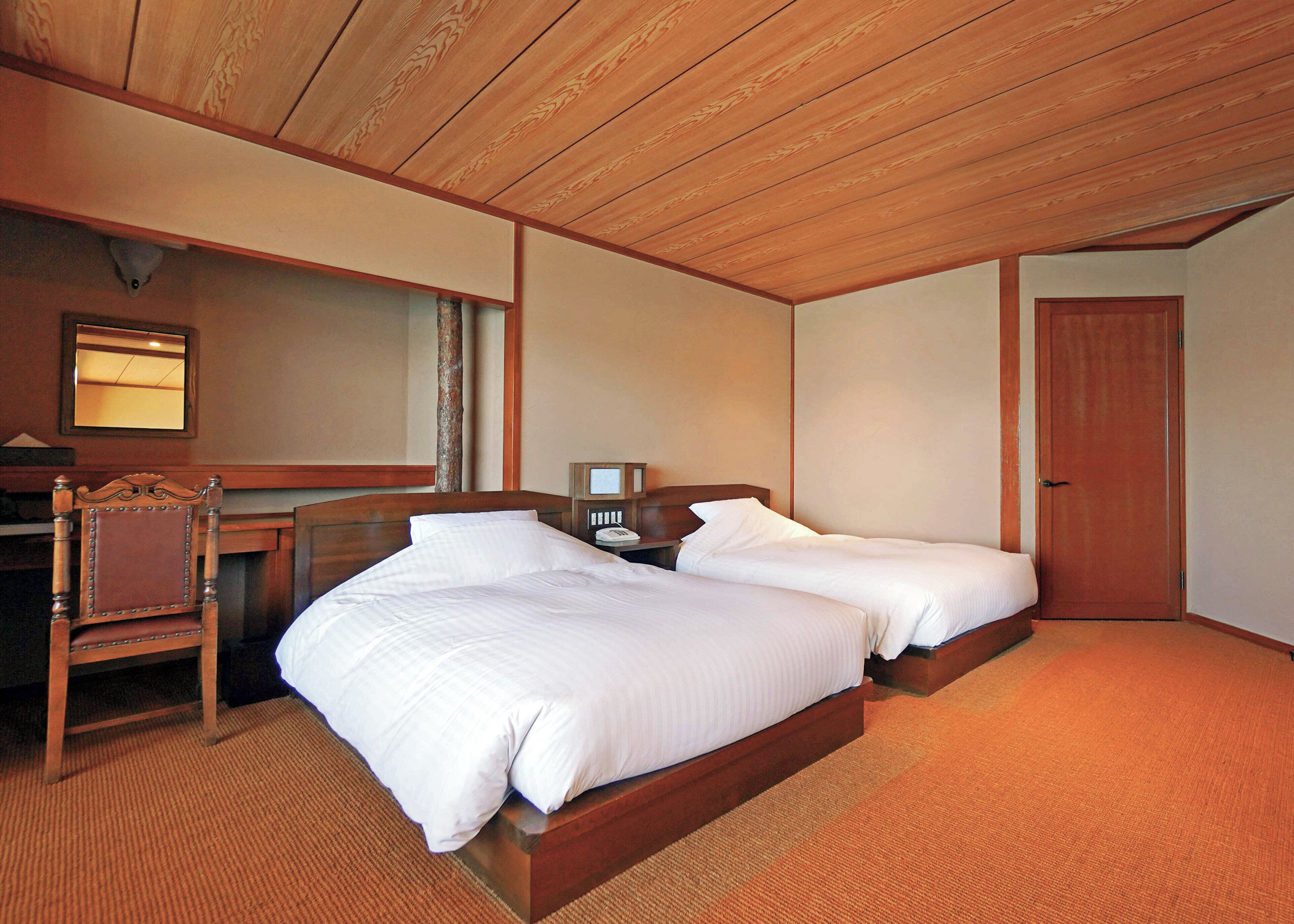 A hotel room with two beds, a wooden desk, a chair, a nightstand, a mirror, and a door.