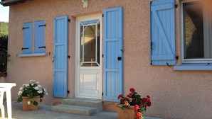 Un bâtiment à l’extérieur recouvert d’un enduit rose, avec des volets bleus, une porte blanche et des marches menant à l’entrée. On y trouve deux jardinières, l’une contenant des fleurs blanches et l’autre, des fleurs rouges.