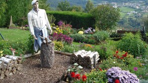 A garden with a statue of a man holding a shovel, surrounded by various flowers and a pile of logs. - GITE upstairs in the owner's house (Haselbourg)