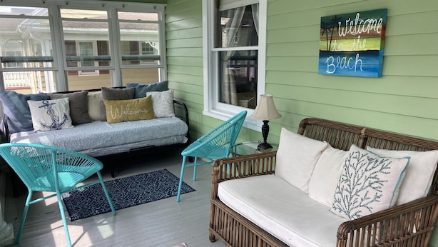 Um quarto aconchegante com parede verde, um sofá com um letreiro que diz 'Bem-vindo à Praia', uma poltrona de vime e uma pequena mesa com um abajur.
