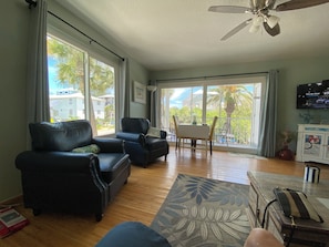 A spacious living room with a large window, a ceiling fan, a dining area, and a television.