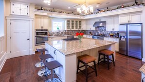 A spacious kitchen with white cabinetry, a granite island with bar stools, stainless steel appliances, and a decorative backsplash.