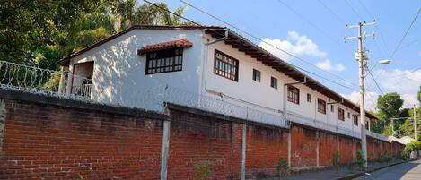 Un edificio de dos pisos con fachada blanca y techo de tejas rojas. Cuenta con un muro de ladrillo con alambre de púas y pequeños árboles plantados a lo largo de la base.