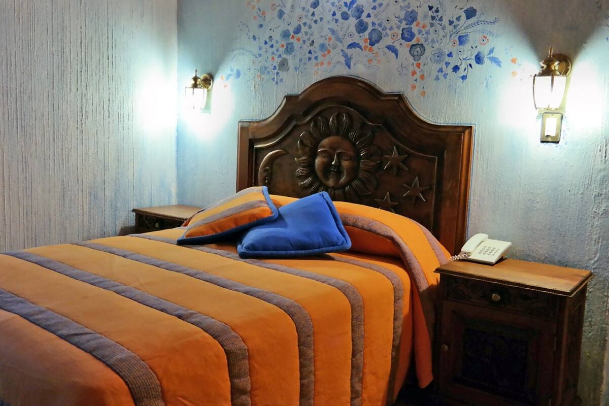 A neatly made bed with striped bedding, a wooden headboard with a carved design, two bedside tables with lamps, and a floral-patterned wall.