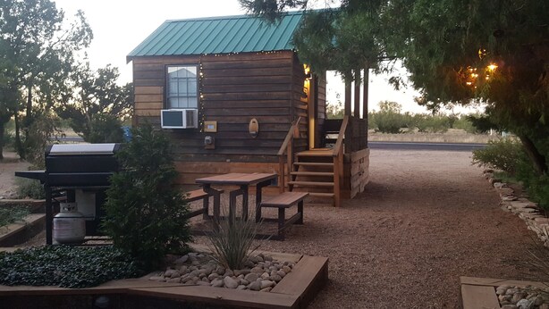 A rustic cabin with a wooden exterior, a small porch, a grill, and a stone pathway leading to the entrance. - The Roadrunner Tiny Cabin at Palo Duro  (Canyon)