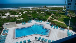 A swimming pool with multiple lounge chairs, a view of the beach, and a building in the background.