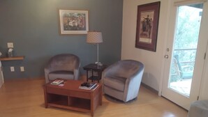 Sala de estar con dos sillones, una mesa de centro y una lámpara. Hay cuadros en la pared y una puerta que da al exterior.