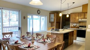A dining area with a wooden table set for four, a kitchen with wooden cabinets and a stove, and a view of the outdoors through a large window. - Bellinger Cabin, wooded getaway near Sisters, Central Oregon (Sisters)