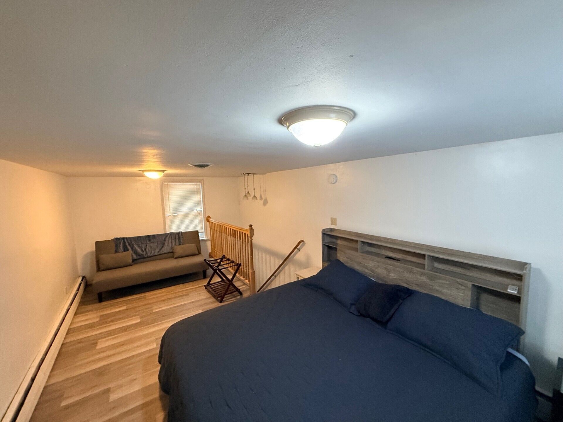 A bedroom with a bed, a sofa, a window with blinds, a wooden floor, and a ceiling light.