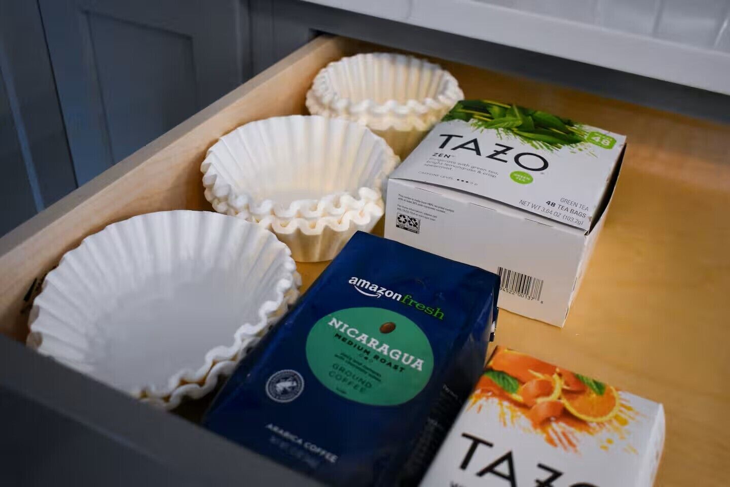Un tiroir contenant des gobelets en papier, une boîte de sachets de thé vert Tazo, un sac de café du Nicaragua, torréfaction moyenne, de la marque Amazon Fresh, et une boîte de sachets de thé Tazo.