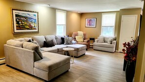 A living room with a sectional sofa, a chaise lounge, and a small round table. There are two windows with blinds, a door, and a framed painting on the wall. - Happy Valley retreat - whole house, large living room, EV charger (Port Matilda)