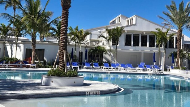 A pool area with palm trees, lounge chairs, and a building with large windows. - Beautiful island escape in beach community (Fort Pierce)