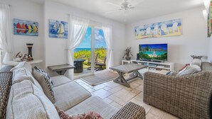 Ein geräumiges Wohnzimmer mit großem Fernseher, bequemen Sitzgelegenheiten und Meerblick durch die Glastür.
