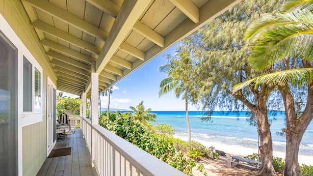 Eine Holzterrasse mit weißer Brüstung, die auf einen Strand mit Palmen und dem Meer blickt.