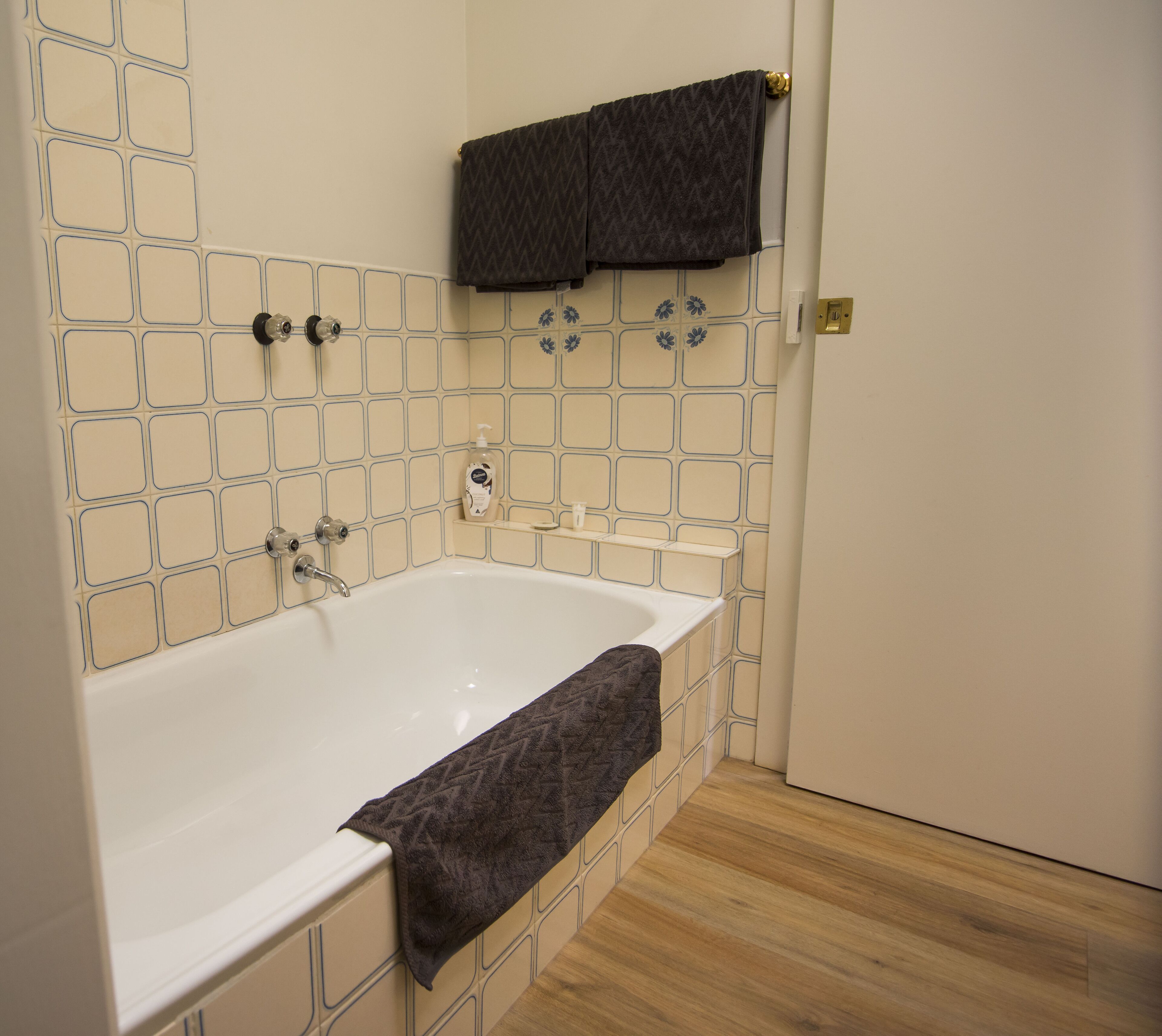 A bathroom with a white bathtub, beige tiled walls, a wooden floor, and a towel hanging on a towel rack.