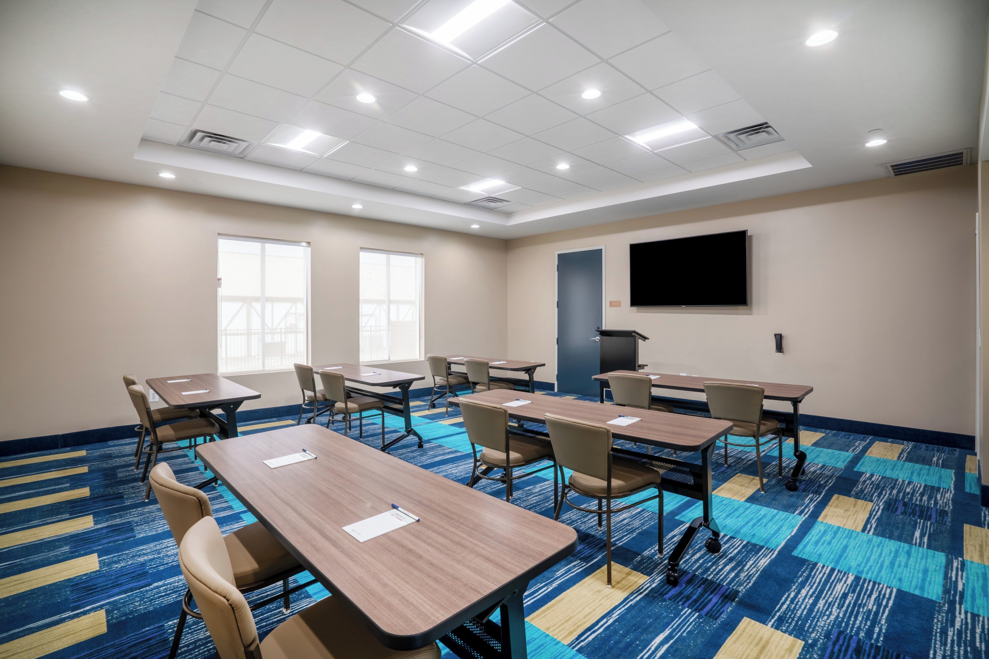 A conference room with tables and chairs, a large flat-screen TV on the wall, and a patterned carpet.