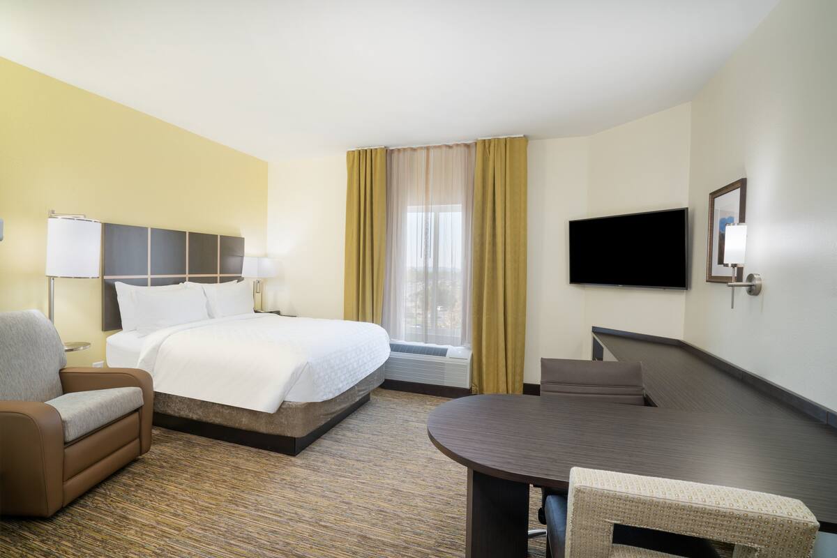 Quarto de hotel com cama, escrivaninha, cadeira, televisão e janela com cortinas.