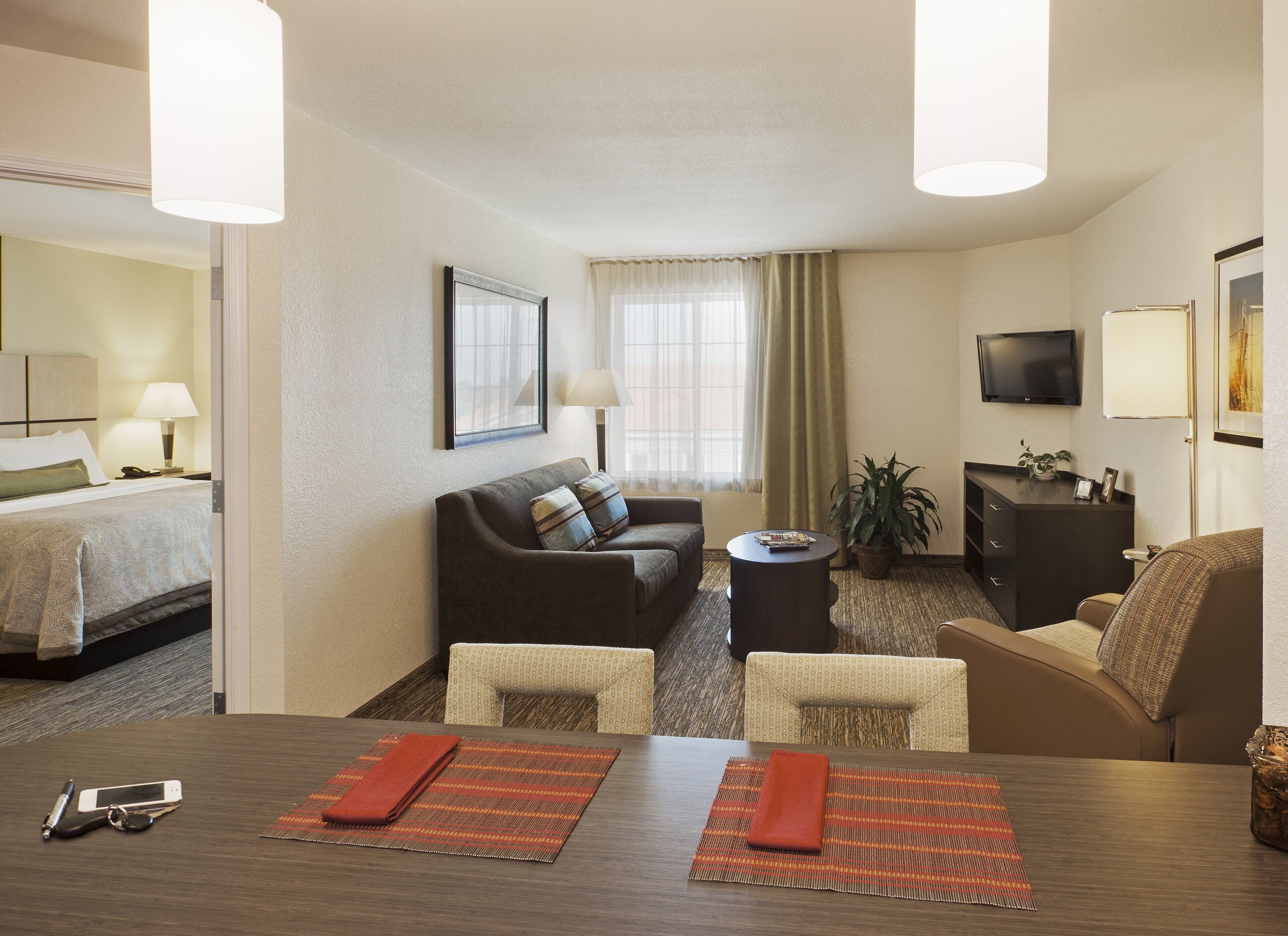 Habitación de hotel con cama, sofá, mesa de comedor con individuales, televisor y planta.