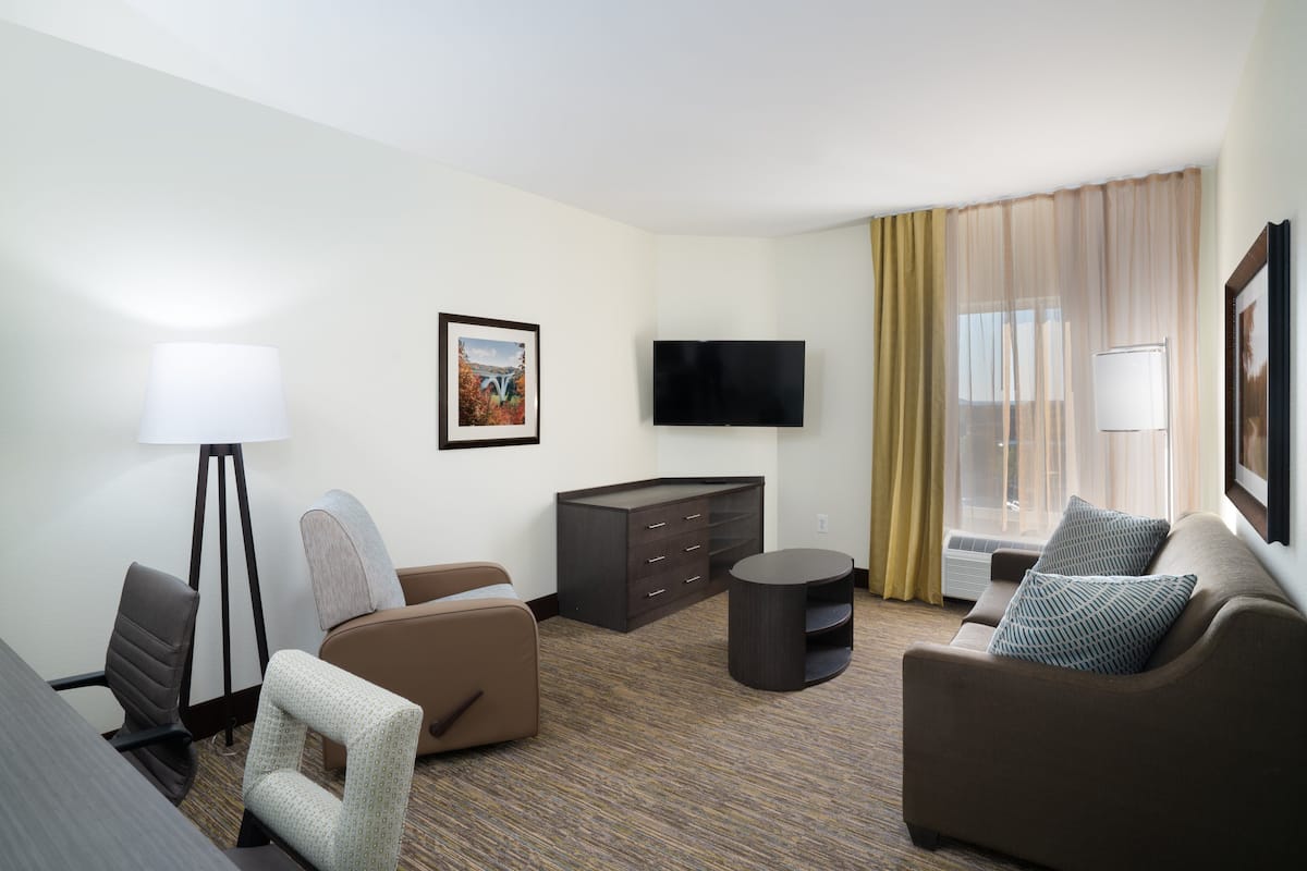 Quarto de hotel com televisão de tela plana, dois sofás, uma mesa pequena, luminária, armário e uma janela grande com cortinas.
