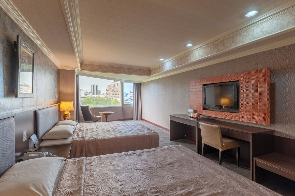 A hotel room with two beds, a desk, a chair, a TV, and a view of the city.