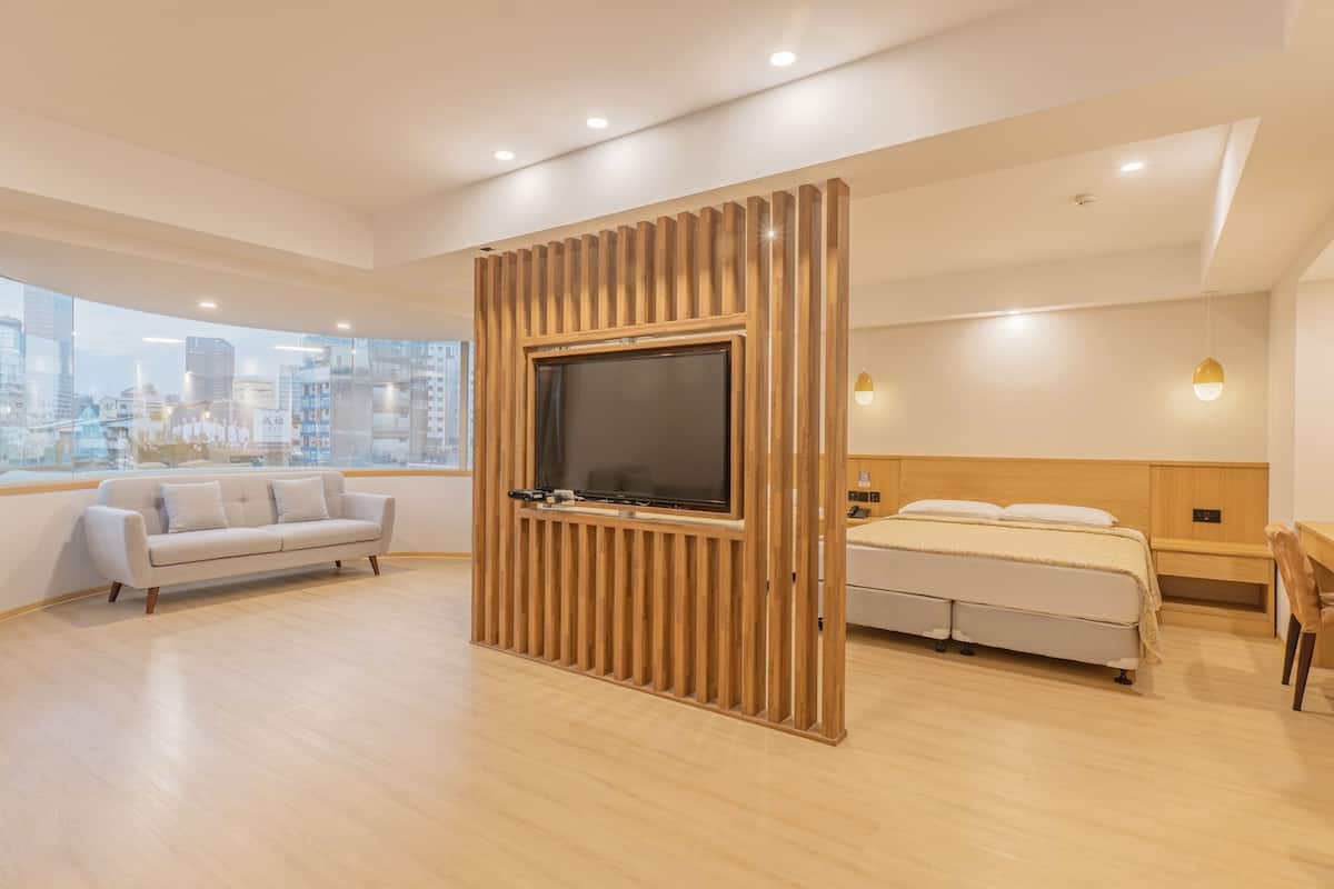 A modern hotel room with a large TV mounted on a wooden panel, a bed with a wooden headboard, a sofa, and a view of the cityscape through large windows.