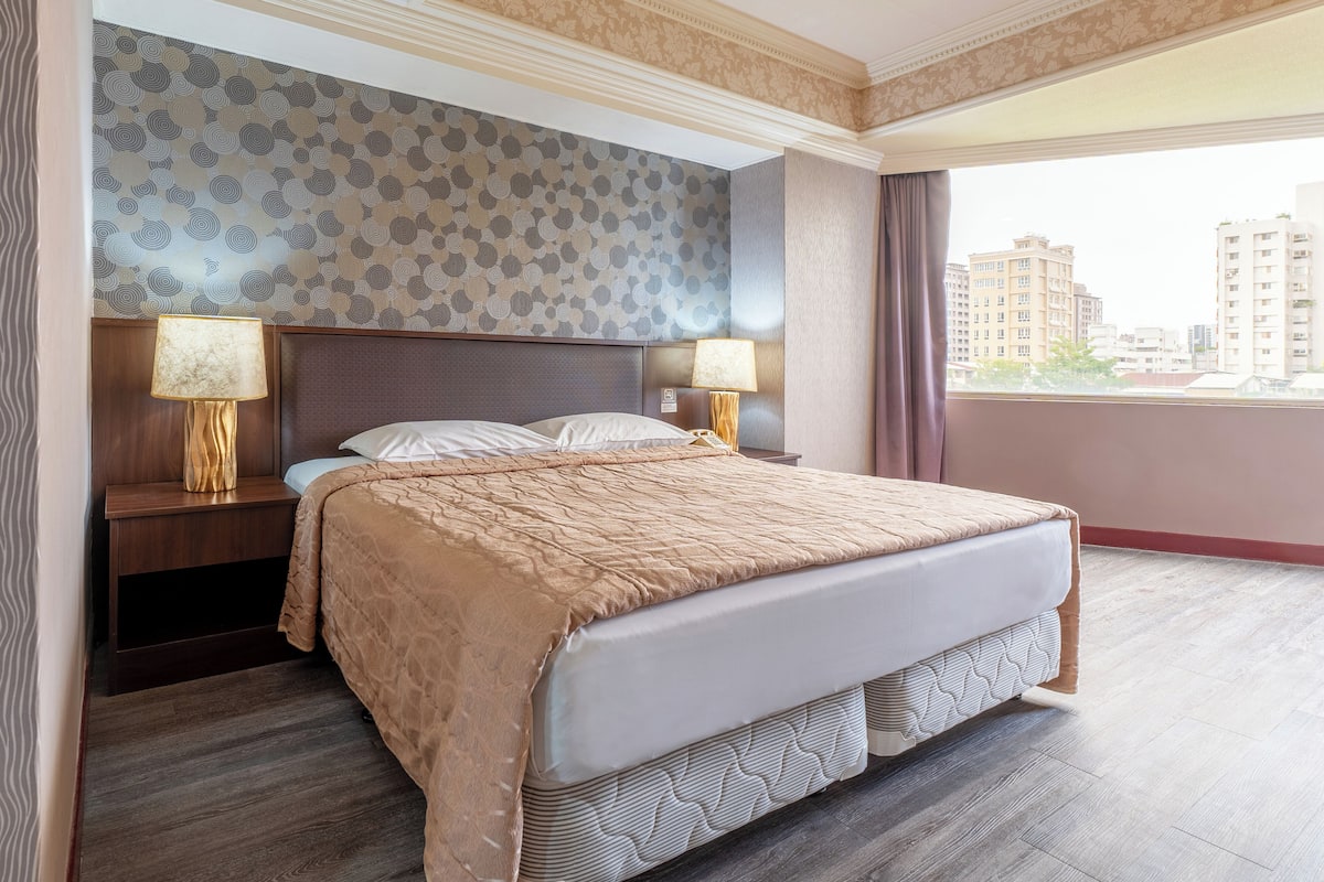 A hotel room with a large bed, two bedside tables with lamps, a patterned wallpaper, and a view of buildings through the window.