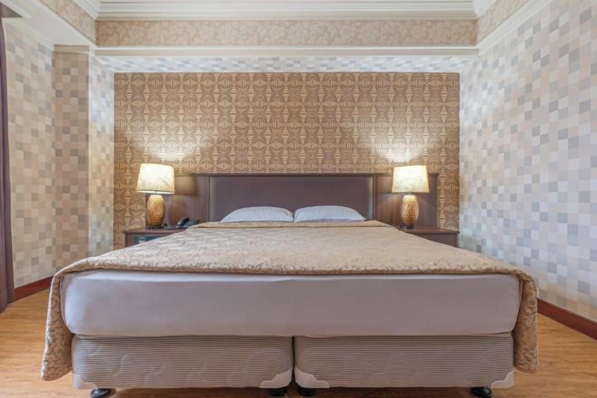 A large bed with a headboard, two bedside lamps, and a patterned wallpaper.
