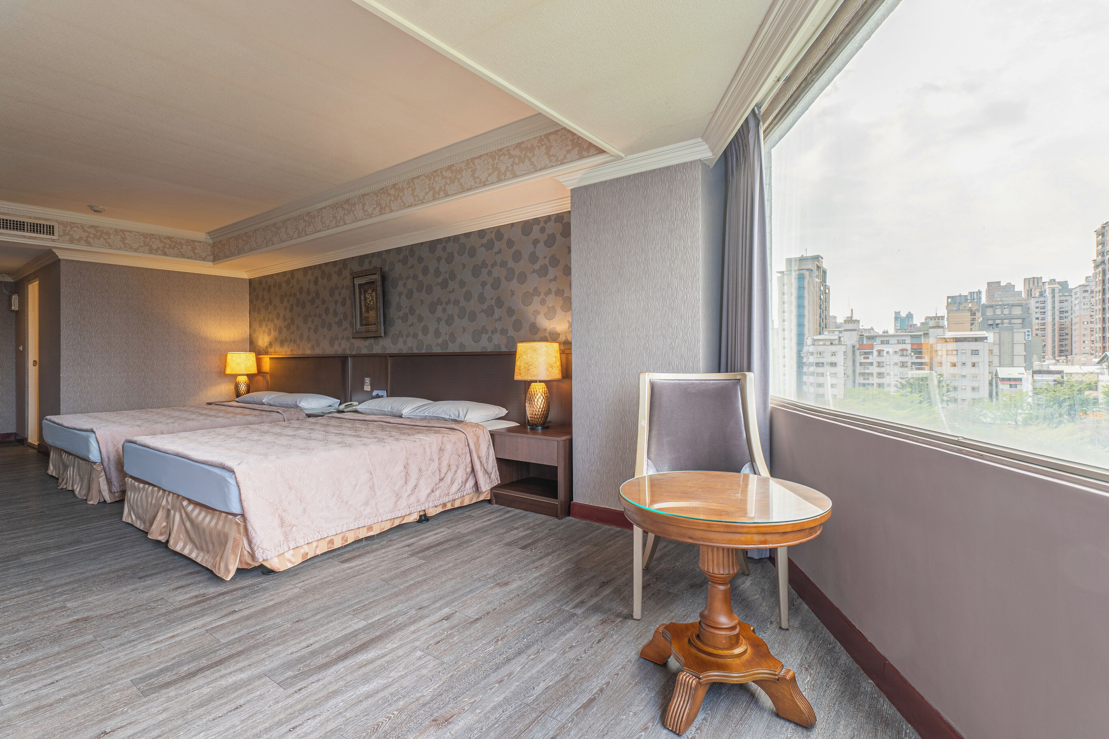 Habitación de hotel con una cama grande, dos mesitas de noche con lámparas, una pequeña mesa redonda y vistas a la ciudad a través de la ventana.