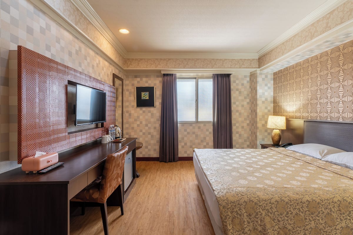 A hotel room with a bed, a desk with a computer, a television, and a window with curtains.