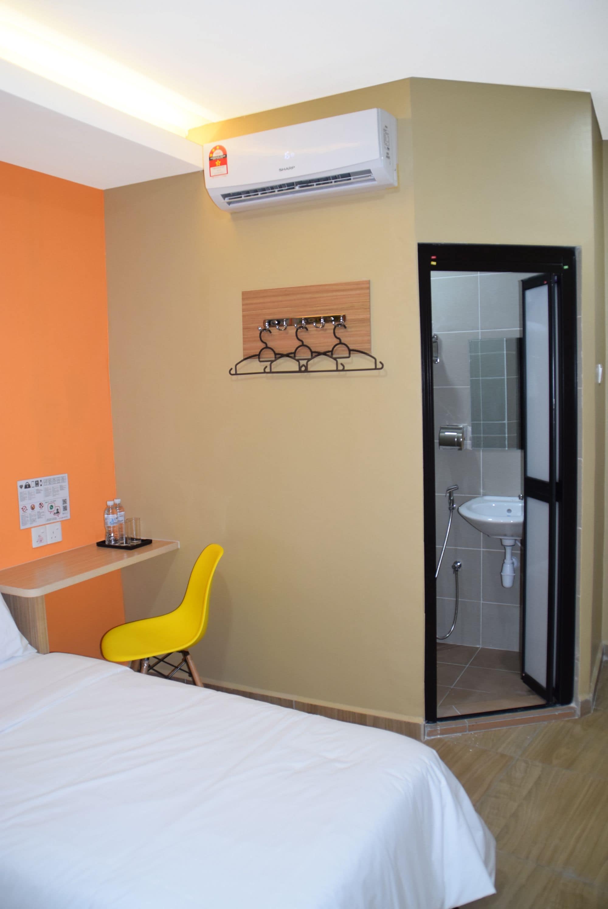 A hotel room with a bed, a desk with a chair, an air conditioner, a bathroom with a sink and shower, and a wall-mounted artwork.