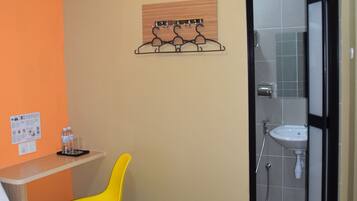 A hotel room with a bed, a desk with a chair, an air conditioner, a bathroom with a sink and shower, and a wall-mounted artwork.