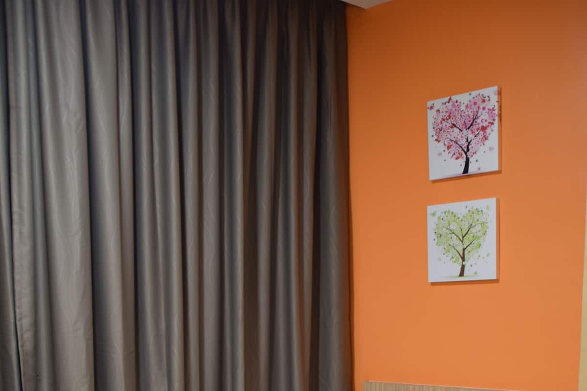 Un lit simple avec une literie blanche, dans une pièce aux murs orange et aux rideaux gris.