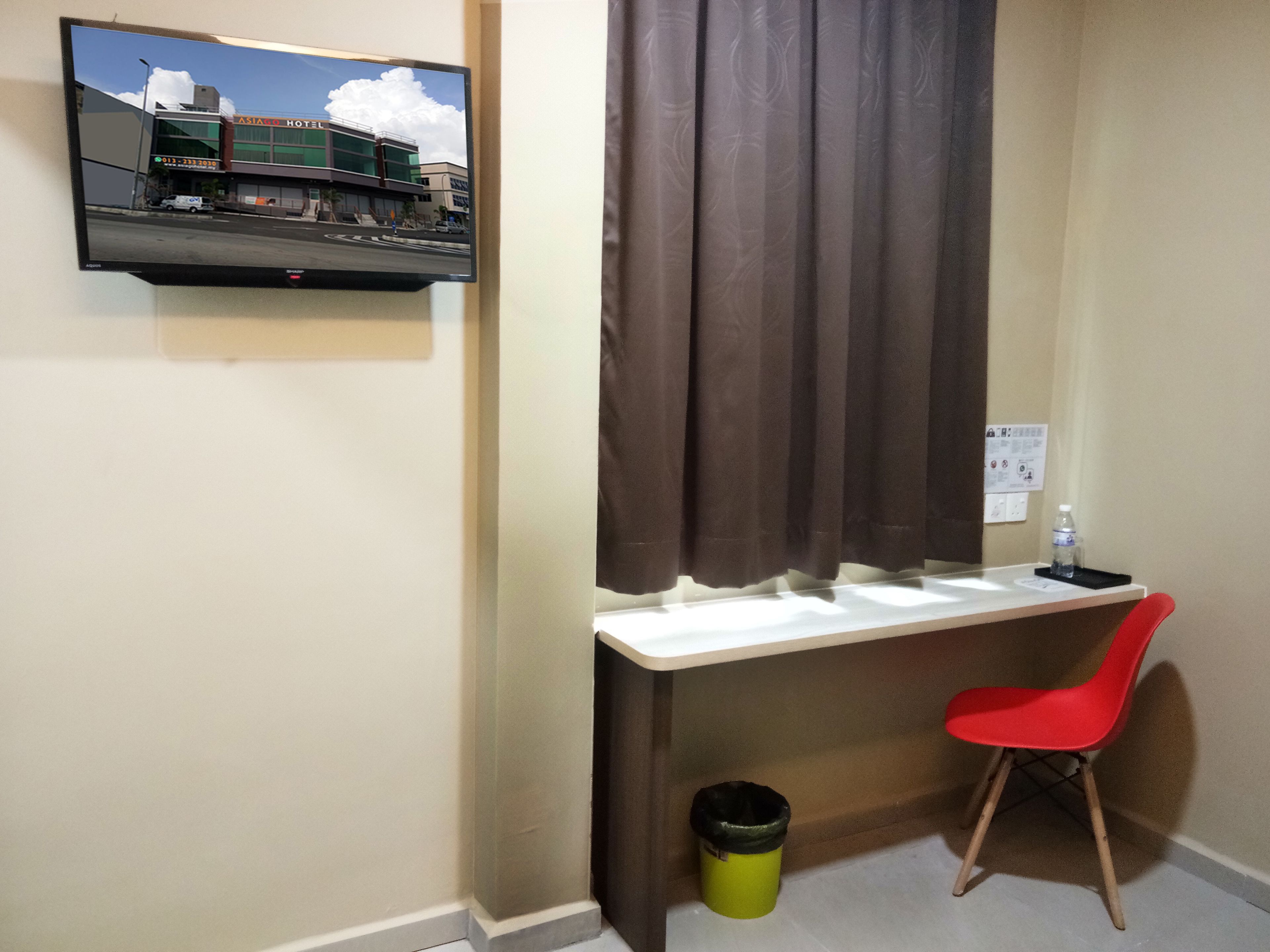 Habitación de hotel con escritorio, una silla roja, televisor montado en la pared y ventana con cortinas.