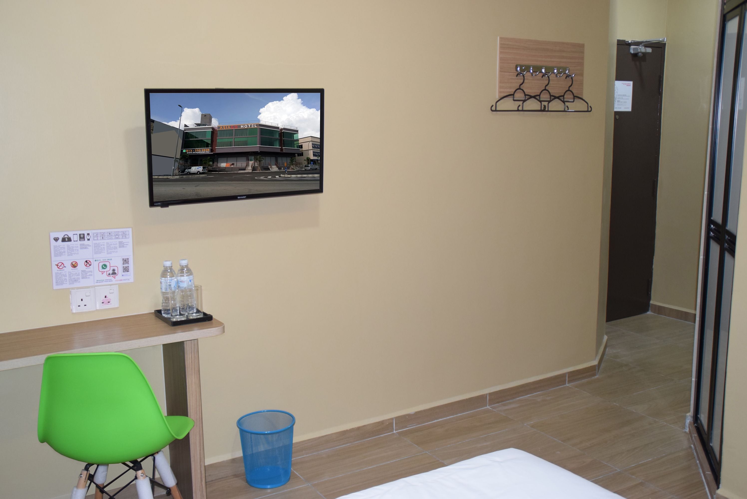 A hotel room with a television mounted on the wall, a desk with a chair, a blue trash bin, and a bed.