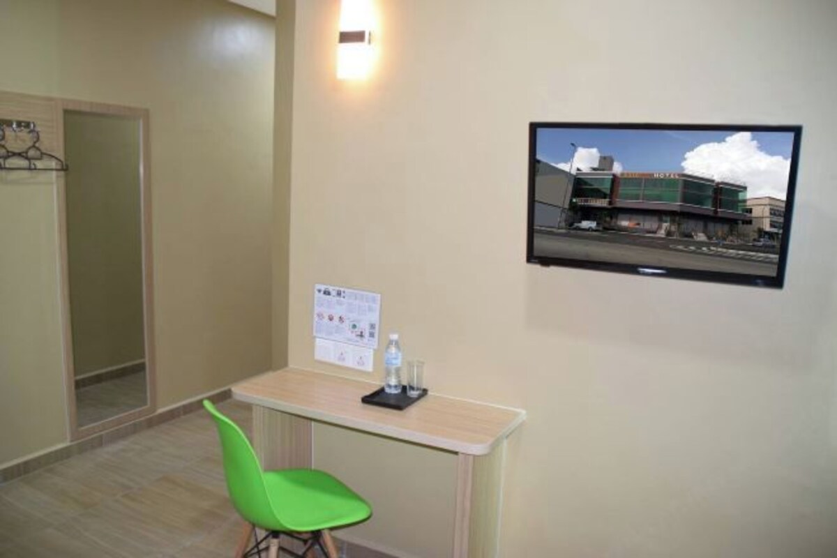 Quarto de hotel com escrivaninha, uma cadeira verde, televisão presa à parede e um quadro de avisos.
