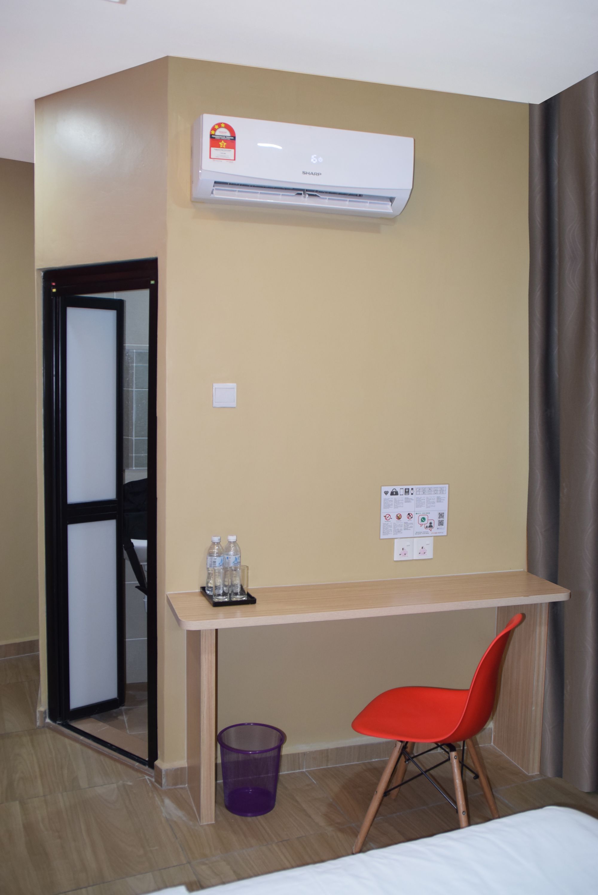 Una habitación de hotel con cama, escritorio con silla, aire acondicionado, un cesto de basura y una puerta.