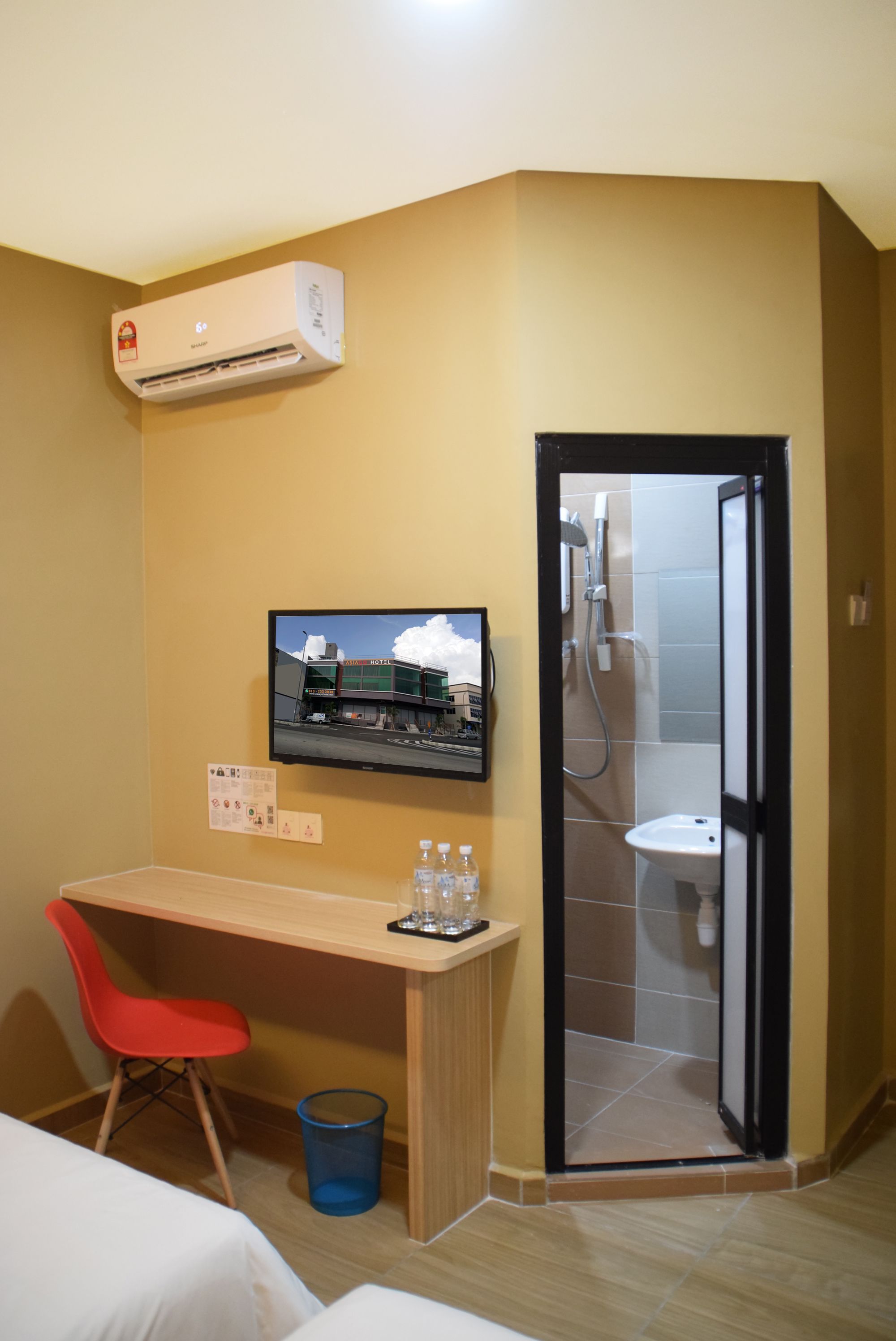Una habitación de hotel con escritorio, una silla roja, televisor montado en la pared y un baño con ducha y lavabo.