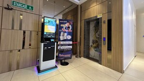 Zona de recepción de un hotel con un quiosco digital, un cartel para la escalera y un ascensor grande con un mural.