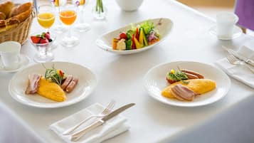 A breakfast setting with plates of eggs, sausages, and a salad, along with juice glasses and a basket of bread.