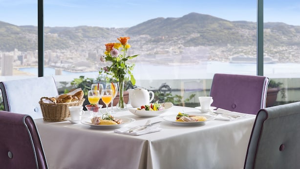 A dining table set with breakfast items, a vase of flowers, and a view of a city and mountains. - Luke Plaza Hotel (Nagasaki)