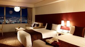 A hotel room with two beds, a sofa, a nightstand, a lamp, and a view of a cityscape at night. - Luke Plaza Hotel (Nagasaki)