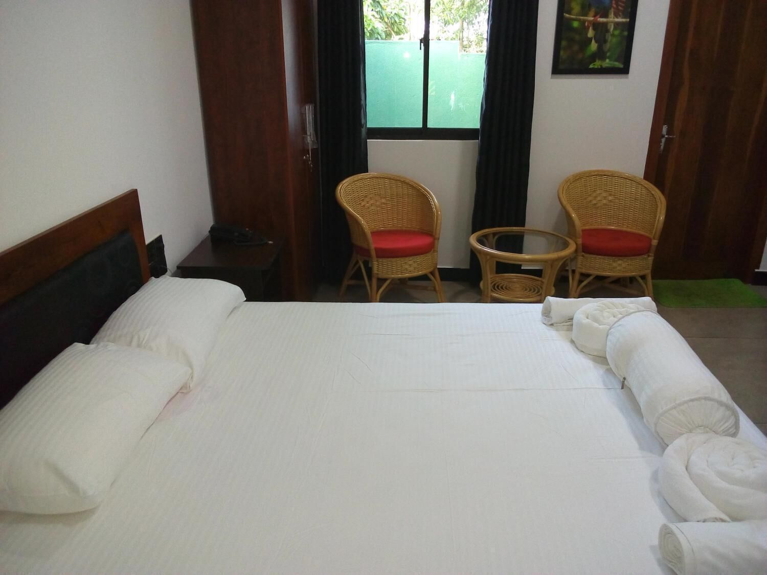 Habitación de hotel con una cama grande, dos sillones de mimbre con cojines rojos, una mesita redonda y una ventana con vistas a la vegetación.