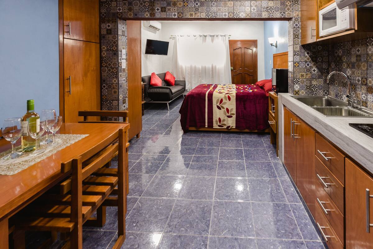 Habitación de hotel compacta con comedor, una cama con cubrecama rojo, televisor montado en la pared y una zona de cocina con microondas.