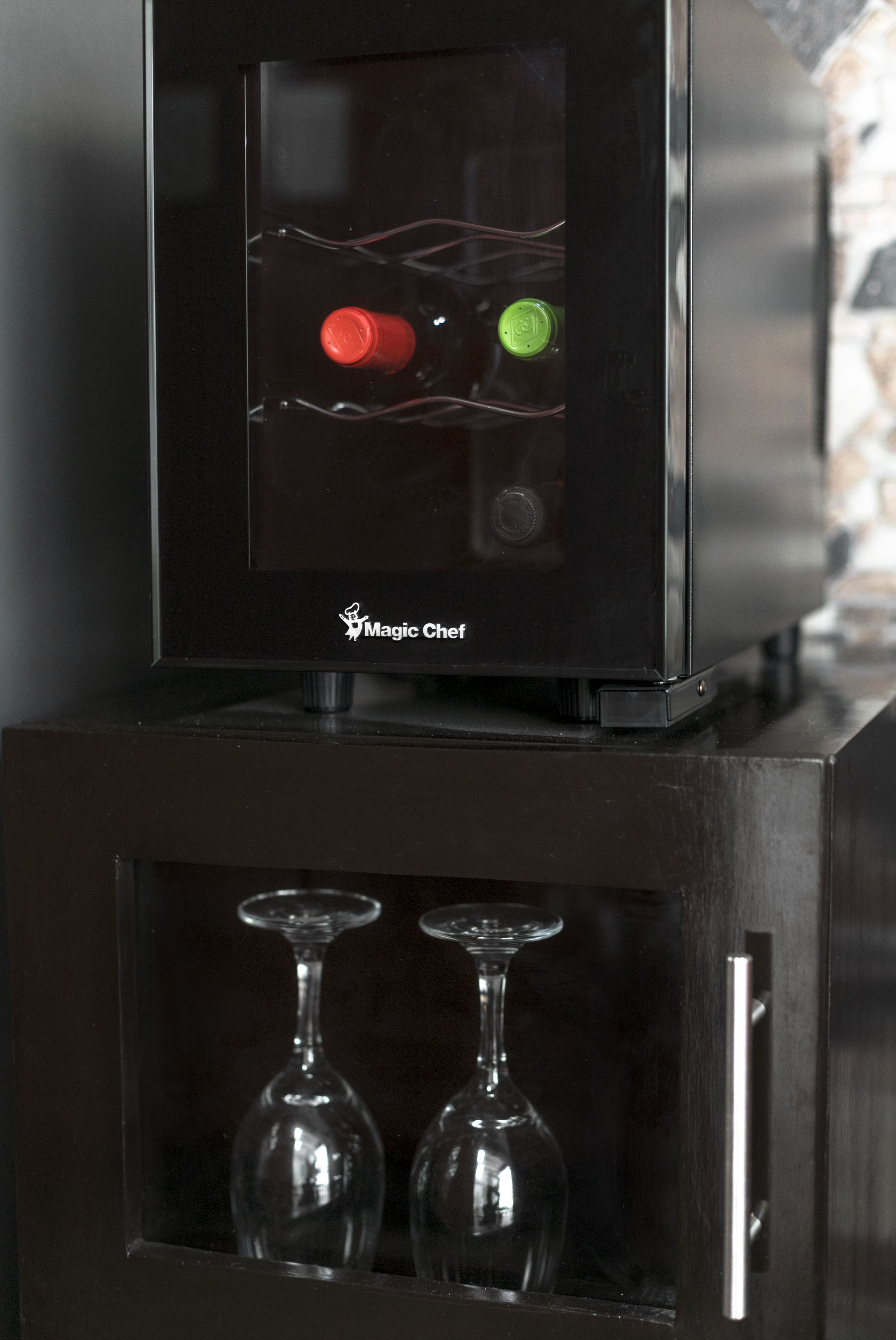 A black minibar with a glass door, containing a red and a green bottle, and two wine glasses.