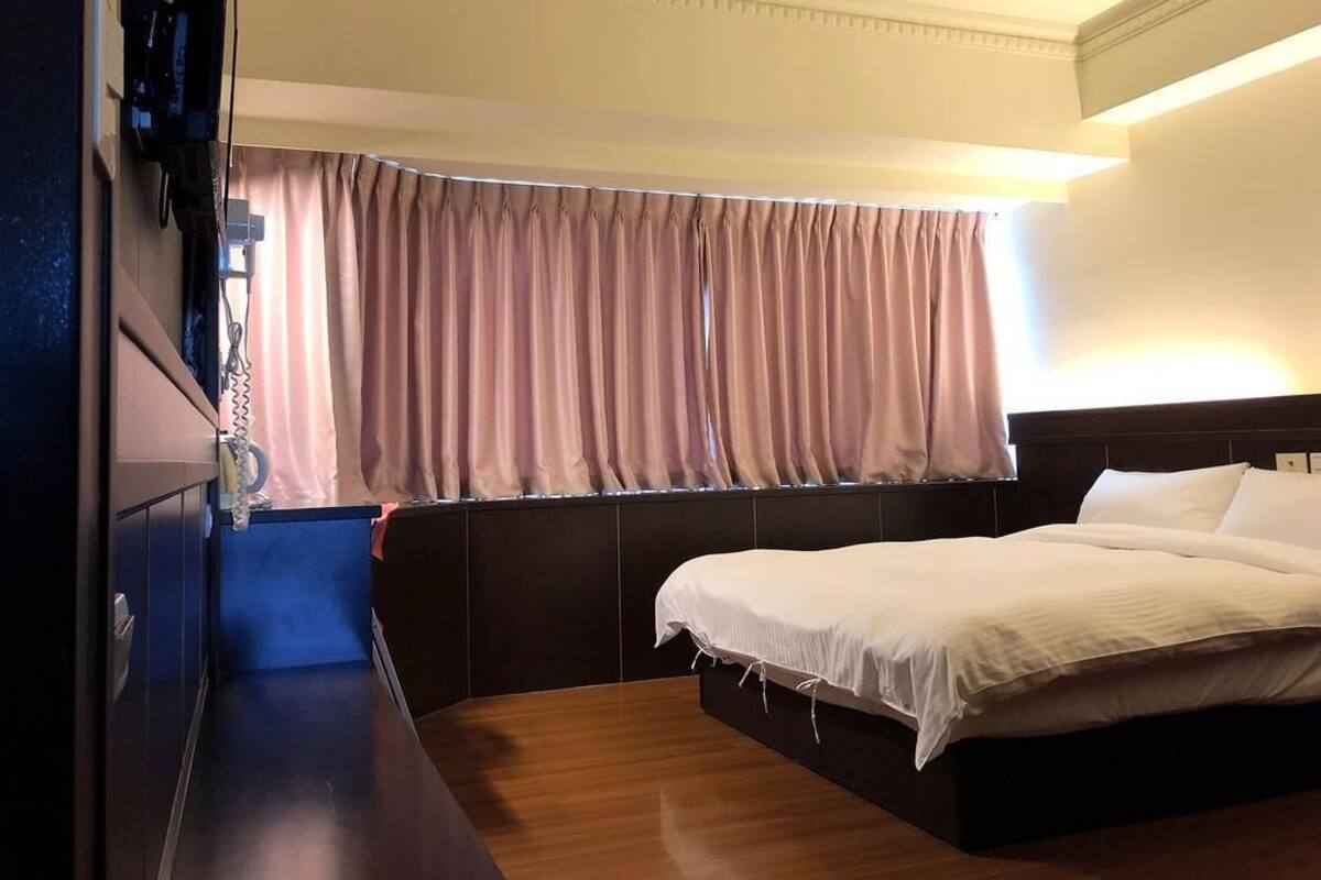 Habitación de hotel con cama, ventana con cortinas, mesita de noche y televisor de pared.