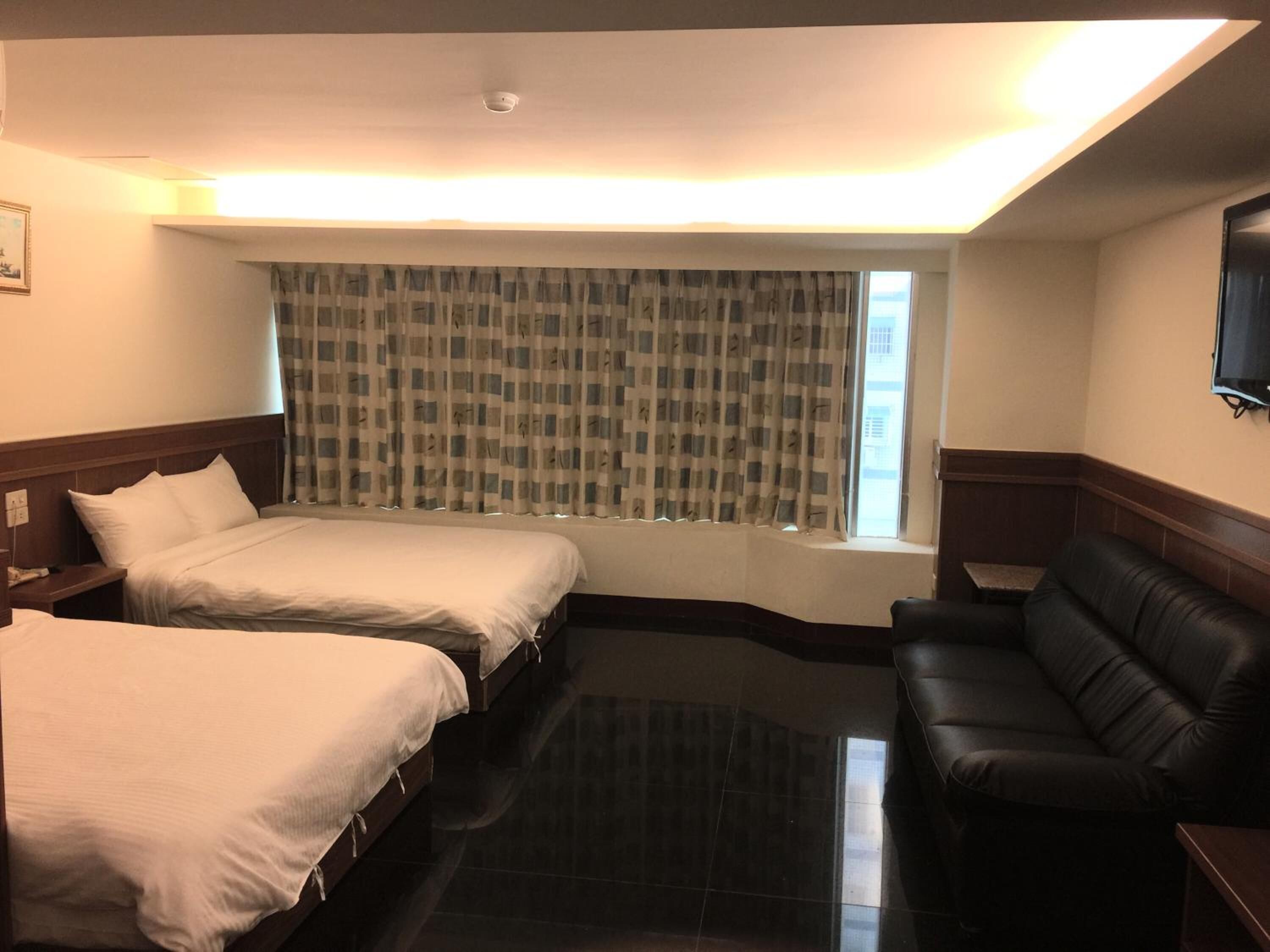 A hotel room with two beds, a black leather sofa, a window with checkered curtains, and a flat-screen TV mounted on the wall.