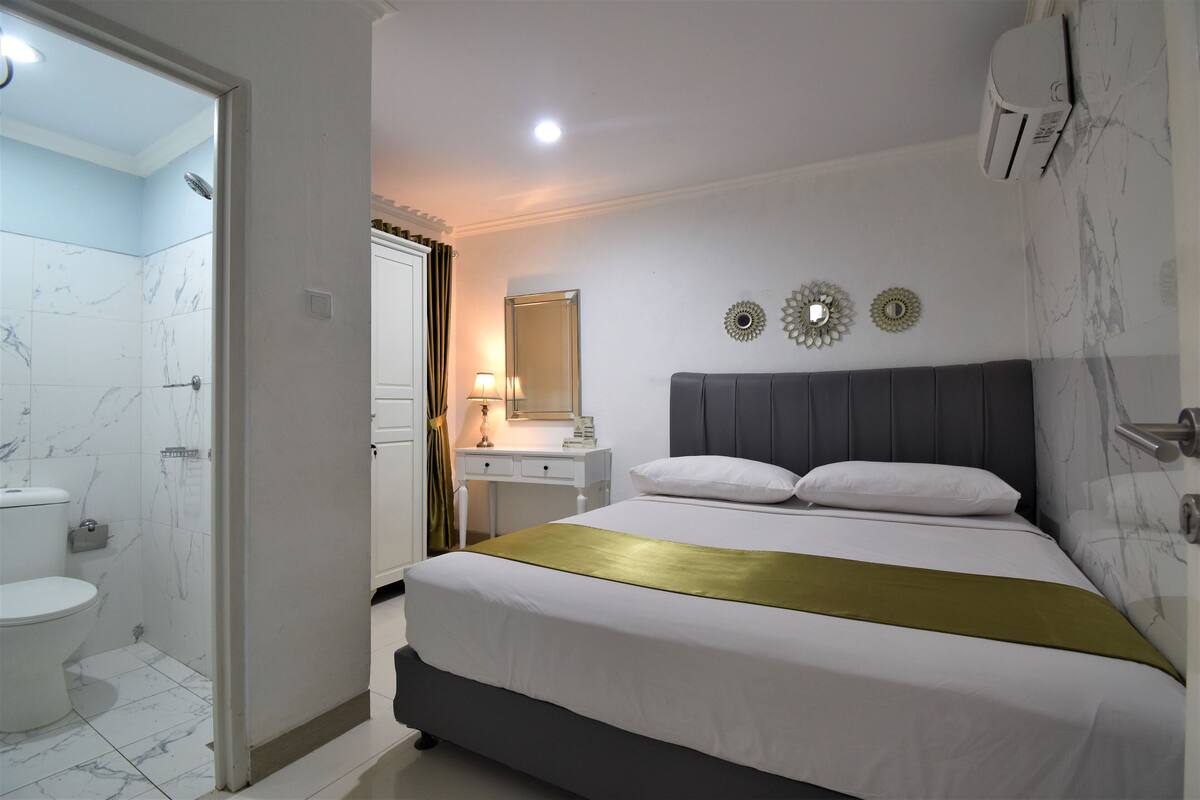 A hotel room with a bed, a bedside table, a mirror, and a bathroom with a toilet and shower.