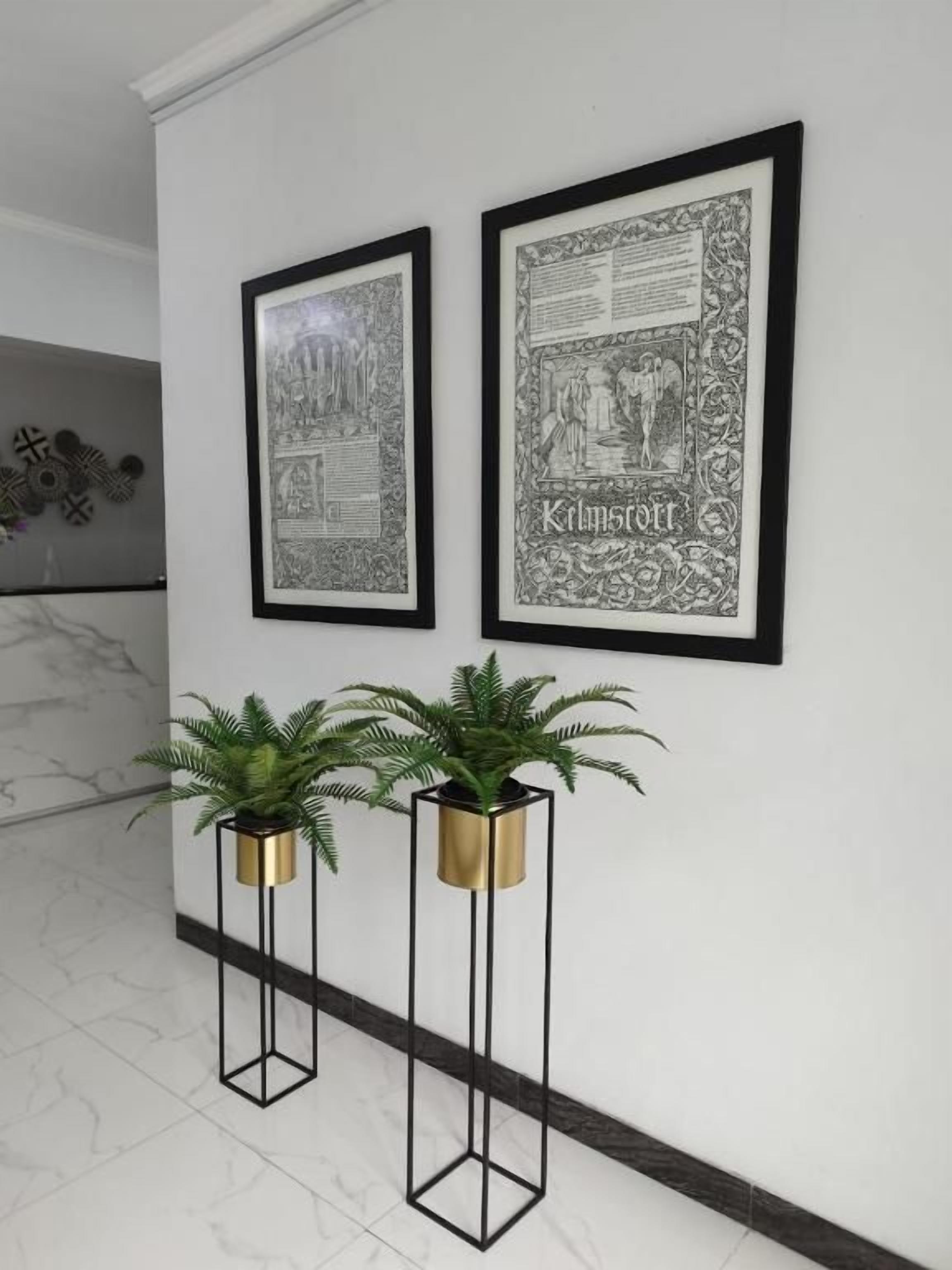 Two framed black and white illustrations on a white wall, flanked by two tall, black metal stands with gold bases, each holding a green plant.